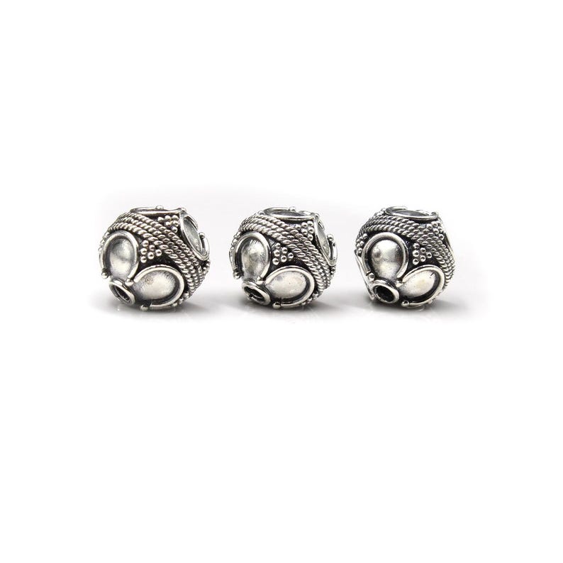 May include: Three silver-colored beads with an intricate design. Each bead features a series of oval and circular shapes, surrounded by a textured, rope-like pattern. The beads are spherical and have a vintage or antique aesthetic.