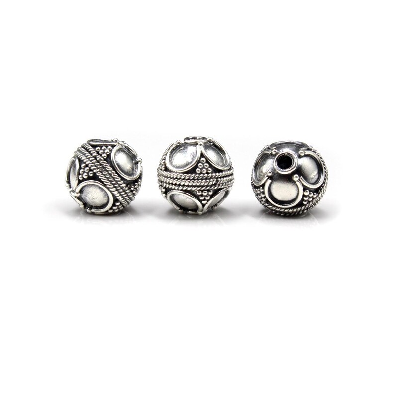 May include: Three silver-colored, spherical beads with intricate designs. Each bead features a central band of twisted rope-like texture, and is adorned with oval and circular patterns. The beads are displayed against a white background.
