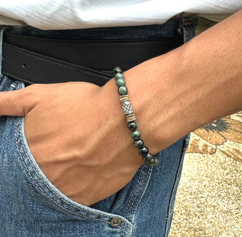 Pu&ograve; includere: Un braccialetto di perline con perline di pietre preziose verde scuro e nere. Il braccialetto ha un elemento decorativo centrale con dettagli metallici argentati e dorati. Il braccialetto &egrave; indossato al polso.