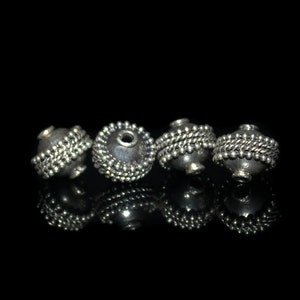 May include: Four silver beads with a textured surface and a small hole in the center. The beads are arranged in a row on a black background.