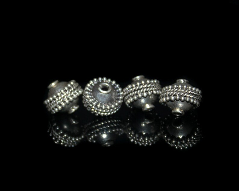 May include: Four silver beads with a textured surface and a small hole in the center. The beads are arranged in a row on a black background.