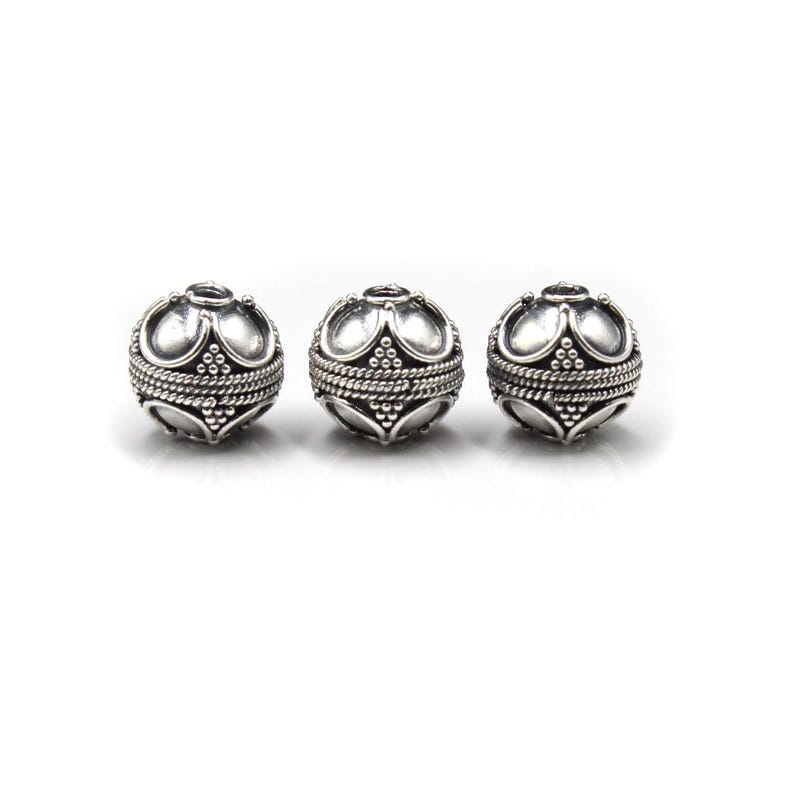 May include: Three silver-colored, spherical beads with an ornate design. Each bead features a crown-like top, a textured band, and a detailed lower section. The beads are displayed against a white background, highlighting their intricate details.