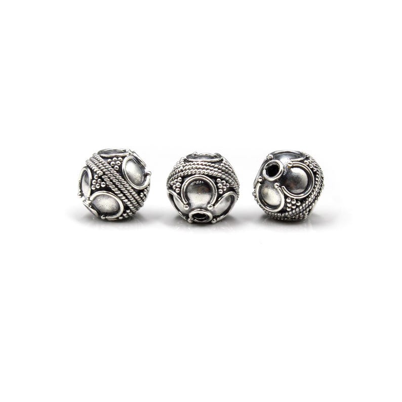 May include: Three silver-colored, spherical beads with intricate designs. Each bead features a pattern of raised dots, curved shapes, and braided rope-like details. The beads are displayed against a plain white background.