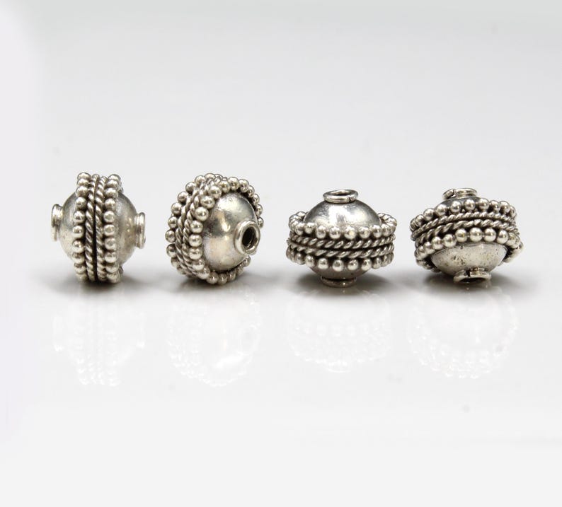 May include: Four silver beads with a textured, beaded design. The beads are round and have a hole in the center for stringing.