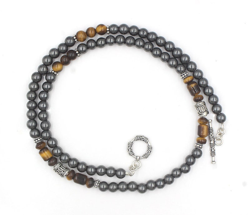 Puede incluir: Un collar de doble cadena con cuentas de hematites grises y cuentas de ojo de tigre. El collar tiene detalles plateados y un cierre plateado.
