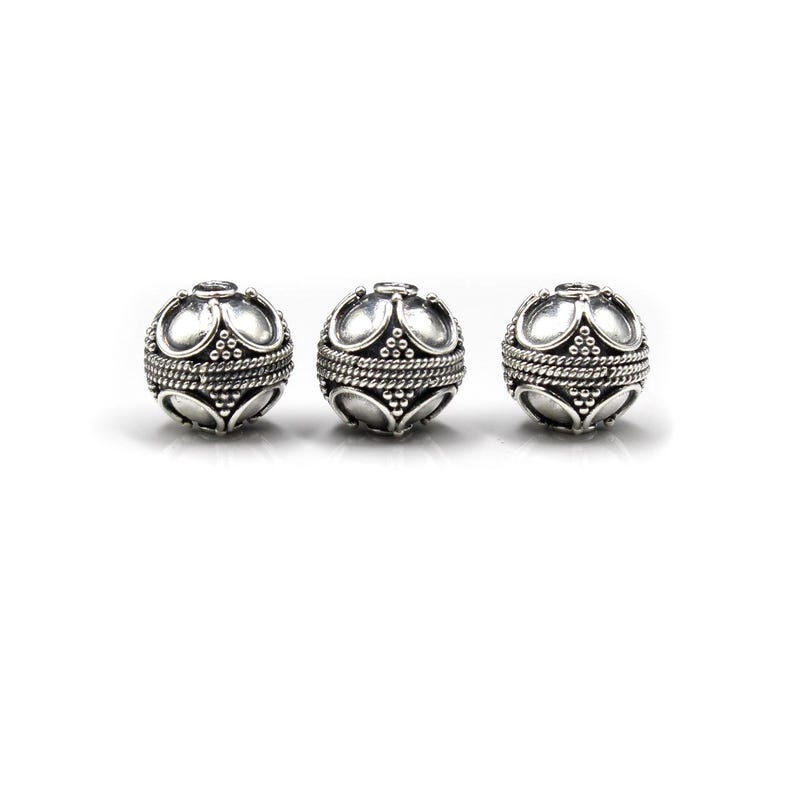 May include: Three silver-colored, spherical beads. Each bead features a decorative pattern of curved shapes and a rope-like band around the center. The beads are displayed against a white background, highlighting their intricate design.