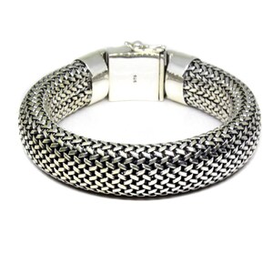 May include: A silver bracelet with a woven chain design. The bracelet is made of a thick, braided chain that is smooth and shiny.