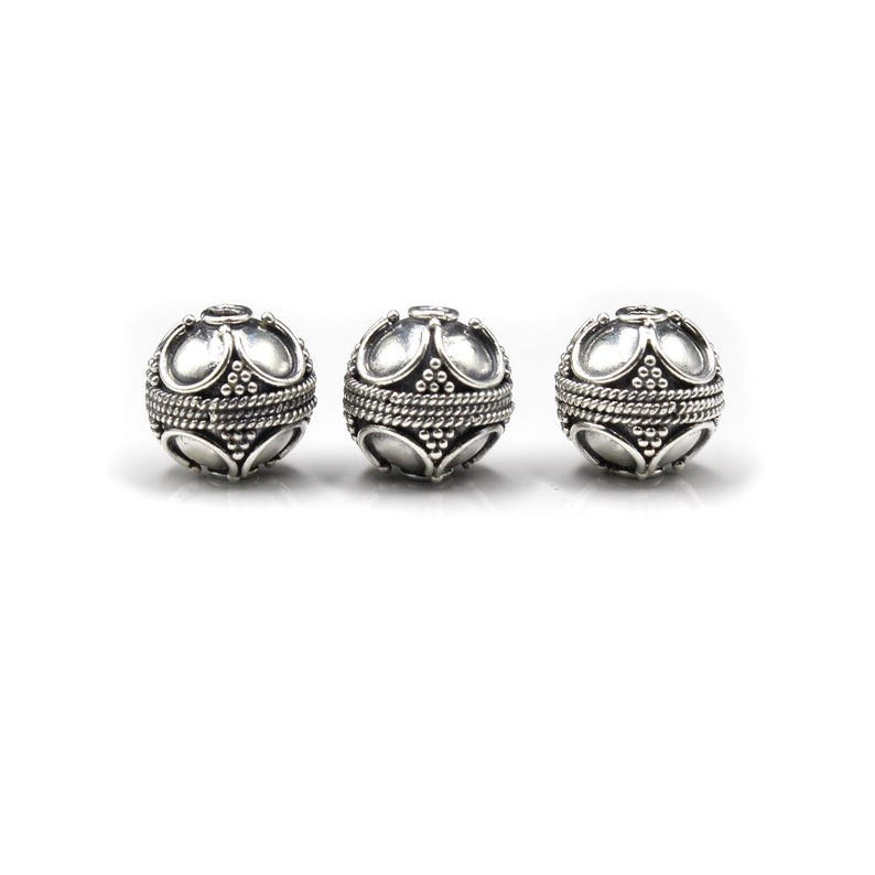 May include: Three silver-colored, round beads with an ornate design. Each bead features a series of curved shapes and a textured band around the center. The beads are displayed against a white background, highlighting their intricate details.