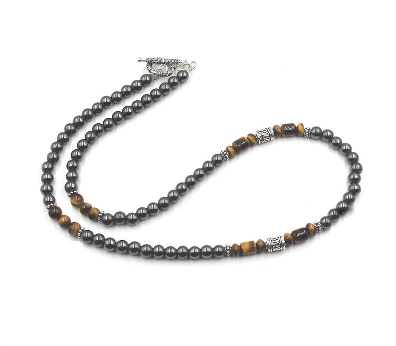 Puede incluir: Un collar de cuentas de hematites negro y ojo de tigre con detalles plateados. El collar tiene un cierre de palanca.