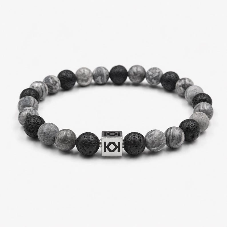 May include: A beaded bracelet featuring a mix of gray, black, and marbled stone beads. A silver cube with a logo is centered on the bracelet. The beads are round and have a natural, textured appearance.