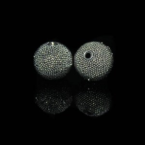 May include: Two round, silver-colored beads with a textured surface, reflecting light. One bead has a hole. The beads are set against a black background, with their reflections visible below.