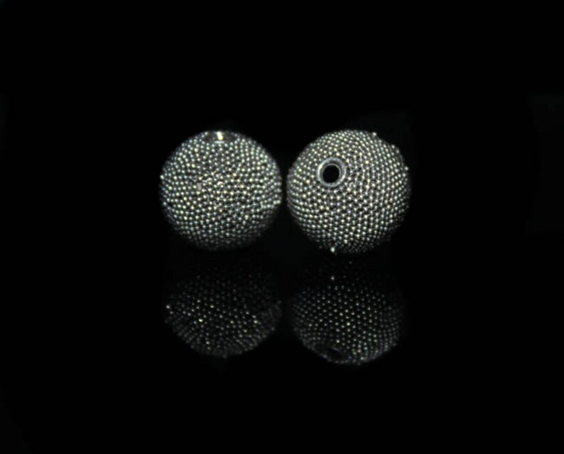 May include: Two round, silver-colored beads with a textured surface, reflecting light. One bead has a hole. The beads are set against a black background, with their reflections visible below.