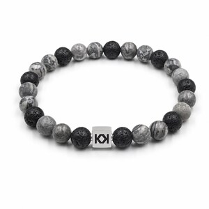 May include: A beaded bracelet featuring a mix of gray and black lava stone beads. A silver square bead with the letters "KI" is centered in the bracelet. The bracelet is on a white background.