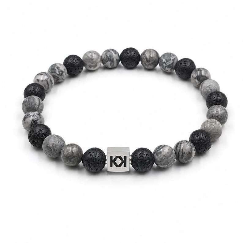 May include: A beaded bracelet featuring a mix of gray and black lava stone beads. A silver square bead with the letters "KI" is centered in the bracelet. The bracelet is on a white background.