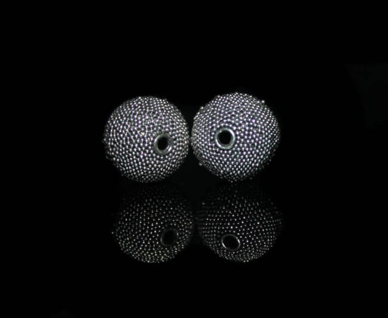 May include: Two silver-colored, spherical beads with a textured surface and a central hole. The beads are displayed against a black background, with their reflections visible below. The beads appear to be made of small, closely packed metallic elements.