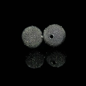 May include: Two silver-colored, spherical earrings with a textured surface, reflecting light. The earrings are set against a black background, with their mirrored reflections visible below. The earrings have a small hole in the center.