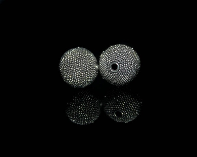 May include: Two silver-colored, spherical earrings with a textured surface, reflecting light. The earrings are set against a black background, with their mirrored reflections visible below. The earrings have a small hole in the center.