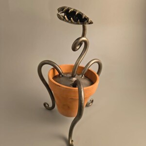 May include: A metal sculpture of a Venus flytrap plant in a terracotta pot. The plant has a detailed, open-mouthed head and curved stems. The pot is a warm orange colour, and the metal is a dark, metallic grey.
