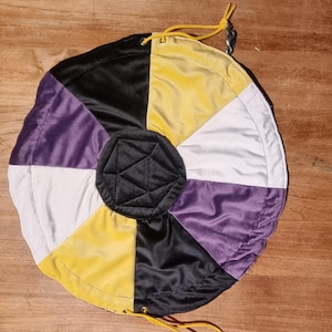 Op de afbeelding: Een ronde, gesegmenteerde tas in de kleuren van de non-binaire pride-vlag: geel, zwart, paars en wit. De tas heeft een trekkoordsluiting met geel koord en een zwart, geometrisch midden. De tas is gemaakt van een glanzend, zijdeachtig materiaal.
