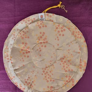 Op de afbeelding: Twee beige, cirkelvormige meditatiekussens met een bloemenpatroon in oranje en geel. De kussens zijn op elkaar gestapeld, met een kleinere bovenop. Een goudkleurige sleutelhanger hangt aan een geel koord dat aan het bovenste kussen is bevestigd. De kussens hebben een label met "Rose Quartz Studio".