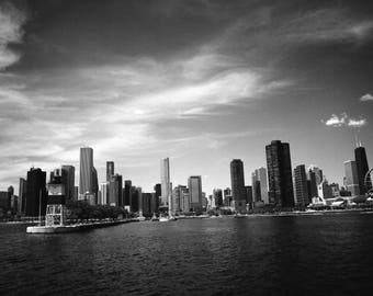 Puede incluir: Fotografía en blanco y negro de un paisaje urbano con edificios altos frente al agua. La imagen incluye una noria y un cielo nublado. El agua es oscura y tranquila.