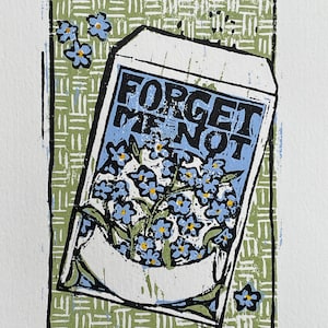 Könnte beinhalten: Ein Druck eines Samentütchens mit dem Text "FORGET ME NOT" in Schwarz auf blauem Grund. Das Tütchen enthält blaue Blumen mit gelben Zentren, vor einem grün-weiß gemusterten Hintergrund. Der Druck ist mit 15/23 nummeriert.
