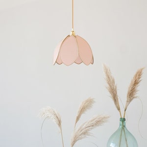 Peut inclure: Un luminaire suspendu en forme de fleur rose est suspendu au plafond contre un mur blanc. Deux vases avec de l'herbe de pampa séchée sont sur une table blanche au premier plan.