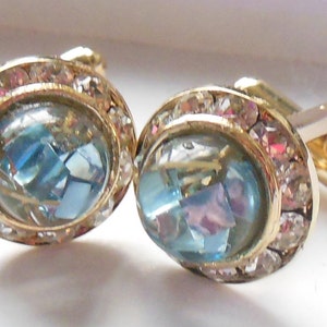 Op de afbeelding: Gouden manchetknopen met een blauwe en iriserende steen omringd door heldere strass steentjes.