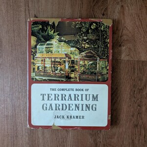 May include: A vintage book titled "The Complete Book of Terrarium Gardening" by Jack Kramer. The cover features a photograph of several terrariums with plants inside, set against a red and white background. The book is on a wooden surface.