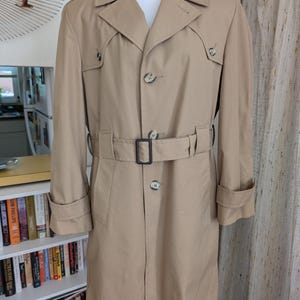 May include: A tan trench coat with a double-breasted front, a belt with a rectangular buckle, and buttoned cuffs. The coat has a classic design with a notched collar and epaulettes. The coat is displayed on a mannequin.
