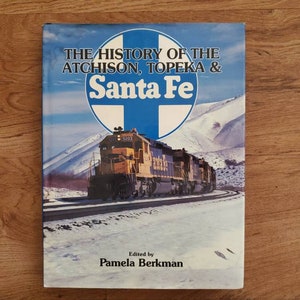 May include: A book titled "The History of the Atchison, Topeka & Santa Fe" featuring a yellow and blue train on a snowy track. The cover has a blue and white logo and the editor's name, Pamela Berkman.