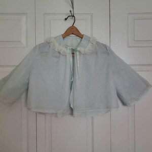 May include: A light blue, sheer bolero jacket with a white ruffled collar and sleeves. The jacket has a tie closure and is hanging on a wooden hanger. The fabric has a subtle polka dot pattern.