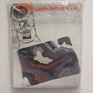 May include: A packaged Latch-a-Rug kit, measuring 24 x 36 inches. The rug design features a mix of colors including black, gray, red, and cream. The packaging includes the brand name and product details.