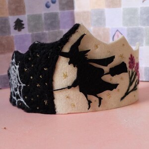May include: A Halloween-themed crown with a split design. One side is black with a spiderweb, the other is cream-colored with a black witch silhouette on a broom, gold stars, and pink flowers. A festive accessory.