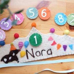 Può includere: Una corona di feltro con un cerchio di feltro verde con il numero 1 in bianco, il nome "Nora" scritto in nero e un animale di feltro marrone con un cappello da festa. La corona è decorata con palloncini, bandierine e pon pon di feltro colorati. Ci sono nove cerchi di feltro con i numeri da 1 a 9 intorno alla corona.