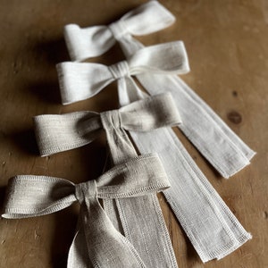 Puede incluir: Cuatro lazos de lino en varios tonos de beige y blanco, dispuestos en fila sobre una superficie de madera. Los lazos están atados con colas largas.