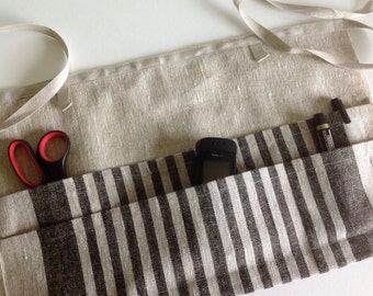 May include: A linen apron with a striped pocket design. The apron is beige with a brown and white striped pocket. A pair of scissors with red handles, a black phone, and two pens are in the pockets. The apron has a beige tie.