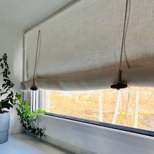 May include: Cream-colored linen window shade with a rolled bottom edge and wooden toggles. The shade hangs in front of a window with a view of trees. A potted plant with green leaves sits on the windowsill.
