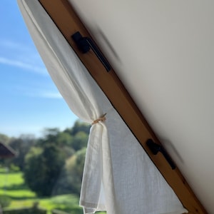 Peut inclure: Un rideau en lin blanc est drapé sur un cadre de fenêtre en bois. Le rideau est maintenu par une pince en bois. La fenêtre offre une vue sur un paysage verdoyant et un ciel bleu.