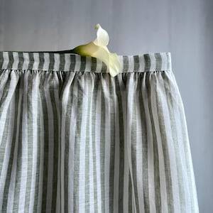 Op de afbeelding: Een rok met verticale strepen in groen- en wittinten. De stof is in de taille gerimpeld. Een witte calla lelie ligt op de bovenrand van de rok en voegt een vleugje elegantie toe. De rok lijkt van linnen te zijn.