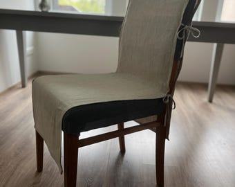 Puede incluir: Una silla de madera con una funda de lino beige. La funda cubre el respaldo y el asiento, asegurada con lazos. La silla tiene patas de madera oscura y un cojín de asiento oscuro. El fondo incluye una mesa y una ventana.