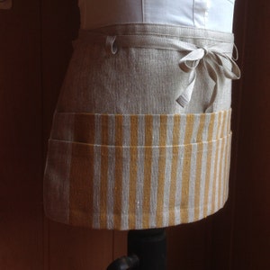 Puede incluir: Un delantal de media cintura de lino con un bolsillo a rayas. El delantal es de color beige natural con un bolsillo a rayas amarillo mostaza y blanco. Tiene un cierre de lazo en la cintura, para cocina o artesan&iacute;a.