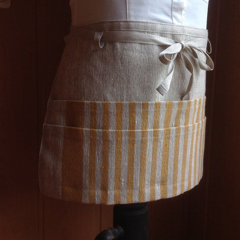 Puede incluir: Un delantal de media cintura de lino con un bolsillo a rayas. El delantal es de color beige natural con un bolsillo a rayas amarillo mostaza y blanco. Tiene un cierre de lazo en la cintura, para cocina o artesan&iacute;a.