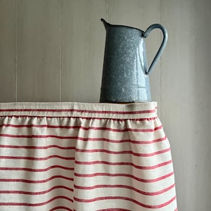 Könnte beinhalten: Ein weiß-rot gestreifter Leinenvorhang mit einem Rüschenrand. Ein grauer Emaille-Krug steht auf einer Holzoberfläche vor dem Vorhang.