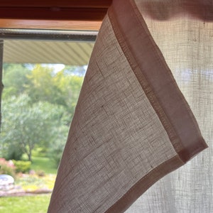 May include: A white linen curtain with a white trim, hanging in a window. The curtain is partially open, revealing a view of a green lawn and trees.