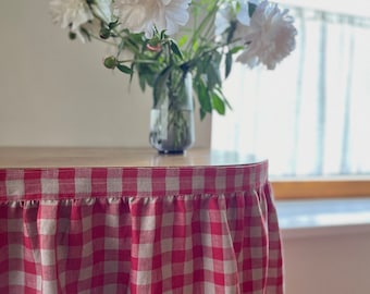May include: Red and white gingham tablecloth with a ruffled edge. The tablecloth is draped over a table with a vase of white flowers in the background.
