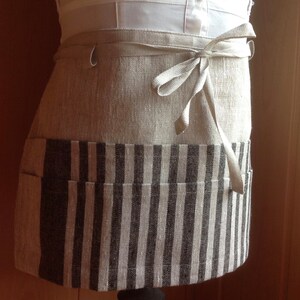 May include: A beige linen half apron with a striped bottom section. The apron has two pockets and a tie-around waist. The striped section features vertical stripes in shades of gray and beige. The apron is tied in a bow.