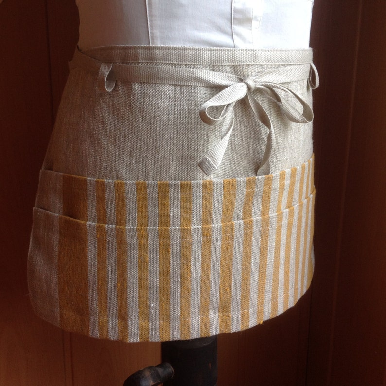 May include: A beige linen half apron with a mustard yellow and white striped pocket. The apron has a tie closure and is displayed on a mannequin. The apron is designed for kitchen use.