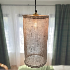 May include: A cylindrical lampshade made of a textured, light brown fabric hangs from a black cord. The lampshade has a gold-coloured fixture at the top and a warm light bulb inside. The background includes green and white curtains.