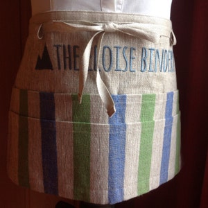 May include: A linen half-apron with a striped pattern in blue and green. The top section features embroidered text that reads "THE MOISE BINDER" above the striped section. The apron has a tie closure and a small pocket.